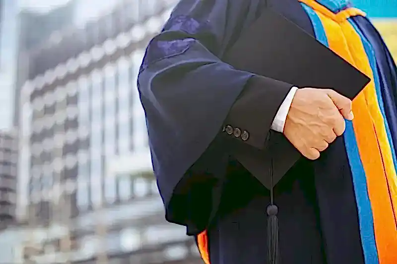 Akademik cübbe giymiş kişi elinde mezuniyet kepi tutuyor – 2025 Mart ve Ekim doçentlik başvuru kriterleri ve ÜAK şartları.