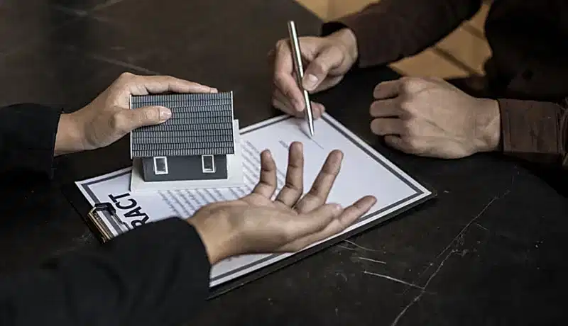 Bir kişinin elinde maket ev tutarken, diğer bir kişinin sözleşme imzaladığı, gayrimenkul hukuku danışmanlığını ve mülk alım satım süreçlerini temsil eden görsel.