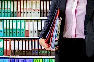 Bir hukuk profesyonelinin (avukat veya ofis çalışanı), arkasında düzenli bir dosya rafı varken elinde dava dosyaları ve önemli belgeler taşıdığı görsel.