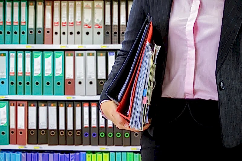 Bir hukuk profesyonelinin (avukat veya ofis çalışanı), arkasında düzenli bir dosya rafı varken elinde dava dosyaları ve önemli belgeler taşıdığı görsel.