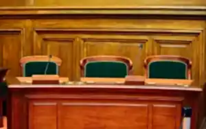Boş bir mahkeme salonunda, hakimin veya jürinin oturduğu ahşap kürsü ve yeşil sandalyelerle, hukuki duruşmaları temsil eden görsel.